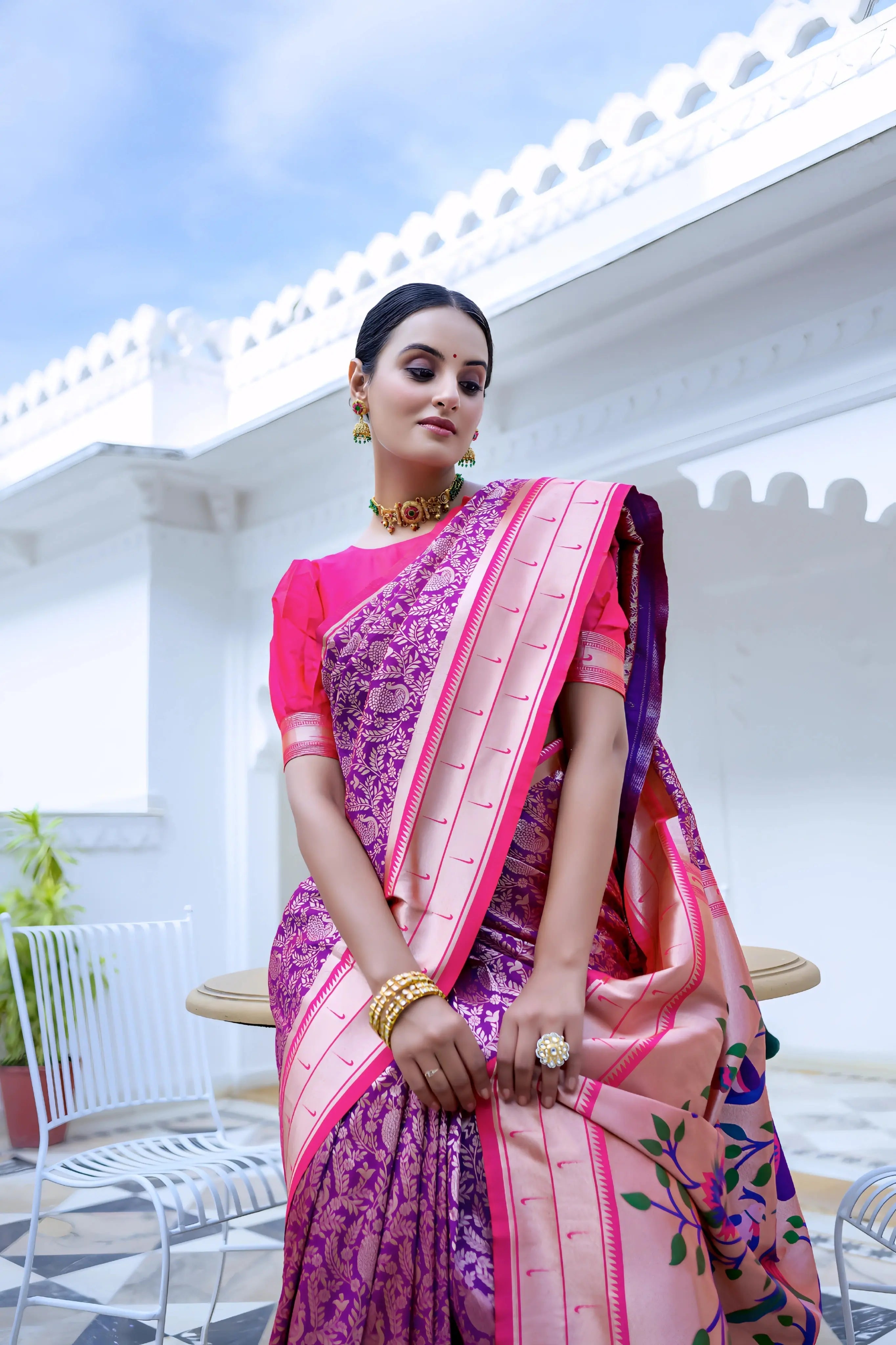 A woman models a vibrant Royal Purple Soft Silk Saree. The saree body features subtle woven patterns. It has a striking light pink contrast border with gold Zari. The pallu is a focal point, featuring rich, artistic Paithani-style print or weaving of colorful bird-on-branch motifs. She wears a fuchsia pink blouse and traditional gold jewelry, posing on an ornate white terrace.